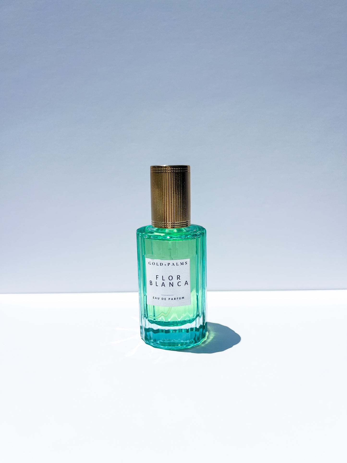 A glass perfume bottle with a gold cap, containing a clear liquid. The label reads 'Flor Blanca Eau de Parfum'.
