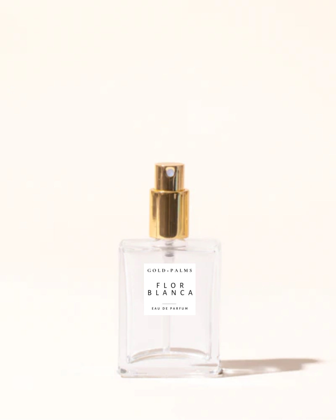 A glass perfume bottle with a gold cap, containing a clear liquid. The label reads 'Flor Blanca Eau de Parfum'.